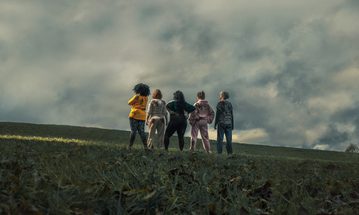 A group of women stand in a line facing away on a grassy hill under a cloudy sky
