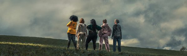 A group of women stand in a line facing away on a grassy hill under a cloudy sky