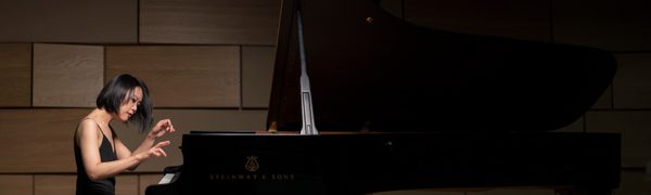 Woman in a floor length black dress sits at a grand piano, and plays energetically, her hair in motion