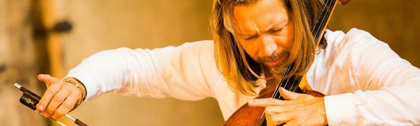 A man with long hair passionately plays the cello in front of a music stand with sheet music