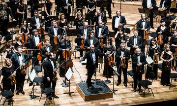 An orchestra dressed in all black stands on stage looking out to the audience