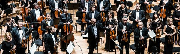 An orchestra dressed in all black stands on stage looking out to the audience
