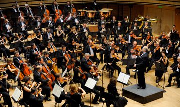 An orchestra dressed all in black sit in a semi-circle formation in a rehearsal room