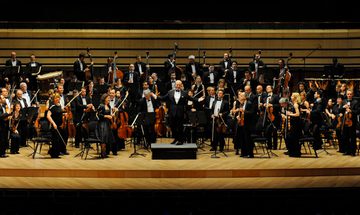An orchestra stands dressed in black on a wooden stage with their conductor smiling with his arms spread in front of them