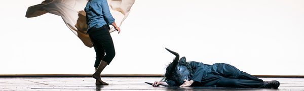 A minotaur lies crumpled on the floor, while a woman stands above him, holding a white cape like a bullfighter. They both are in front of a stark white background.