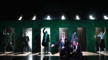 A man keels on a misty stage with a series of doors in the background with his arms outstretched