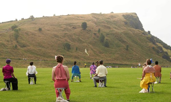 People in colourful clothing sit on stools spaced out in front of a cliff