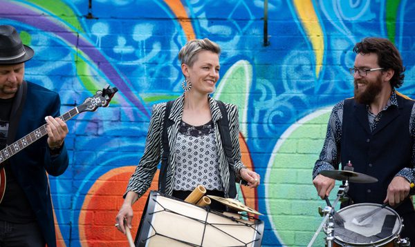 Three musicians perform with drums and banjo in front of a colourful mural