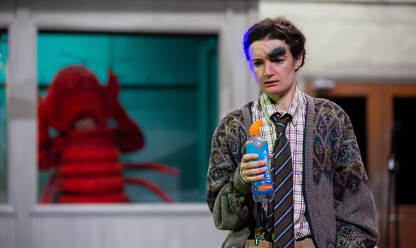 A woman with one large eyebrow holds a bottle of lucozade, as a woman in a lobster costume stands in the background