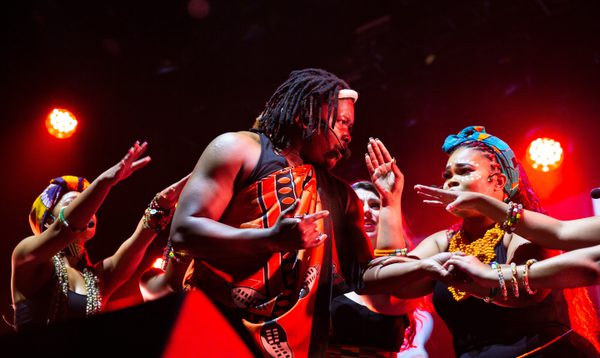 Four performers gesture to each other, wearing brightly coloured African headwraps, wrappers and jewellery