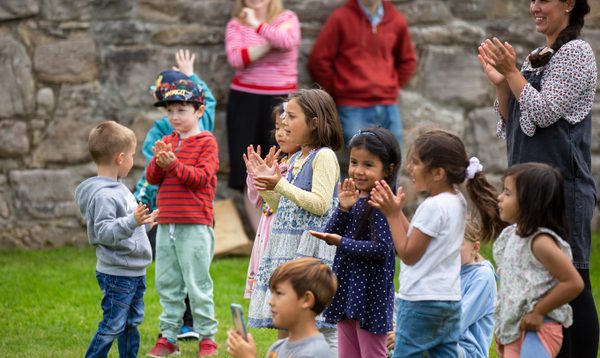 A group of young children applaud, standing on grass in front of a stone wall
