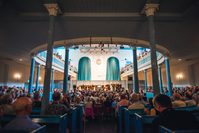 The interior of The Queen's Hall, with audience members sitting in church pews, below blue columns
