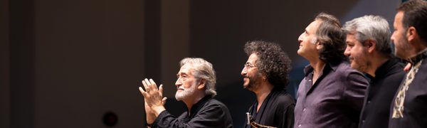 A group of musicians stand on stage smiling at the audience. A man on the end with grey hair and a grey beard applauds the audience.