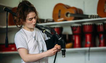 A woman in a white t-shirt with brown reddish hair piled on top of her head plays the Scottish smallpipes