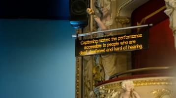 A black screen above an ornate box next to the proscenium arch of a theatre is illuminated with text explaining captioning.