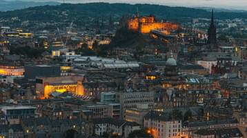 The city of Edinburgh pictured from above at dusk, with the castle and some tenement windows illuminated by amber lights.