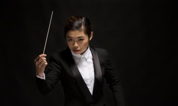 The conductor shown holding her baton and looking up at the camera, dressed in a white shirt and black tuxedo jacket.