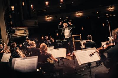 Conductor Thomas Sondergard stands in front of the orchestra with baton in the air