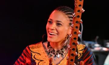 Sona Jobarteh, wearing elaborate jewellery and bright clothing, poses with the musical instrument, the kora
