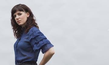 Anna Meredith pictured standing in profile with her head turned towards the camera, wearing jeans and a blue shirt with ruffled sleeves.