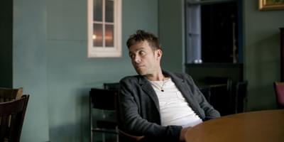 Damon Albarn pictured wearing a white shirt and grey jacket, leaning back in a chair and looking out to his right
