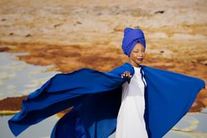Fatoumata Diawara stands in a desert landscape, wearing a blue cape that billows around her.