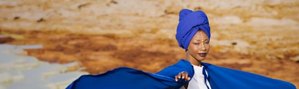 Fatoumata Diawara stands in a desert landscape, wearing a blue cape that billows around her.
