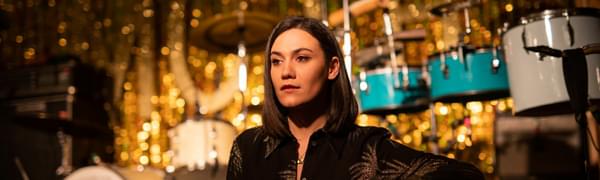 Nadine Shah sits in front of a stage with drum kits and a gold backdrop.