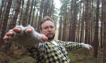 Richard Dawson stands in a forest, reaching towards the camera, wearing fingerless gloves and a plaid shirt.