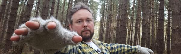 Richard Dawson stands in a forest, reaching towards the camera, wearing fingerless gloves and a plaid shirt.