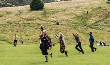 A row of smiling people perform a dance in a field, with Arthur's Seat in the background.