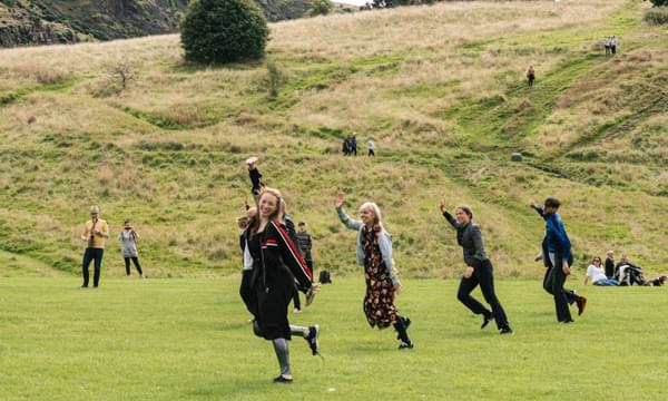 A row of smiling people perform a dance in a field, with Arthur's Seat in the background.