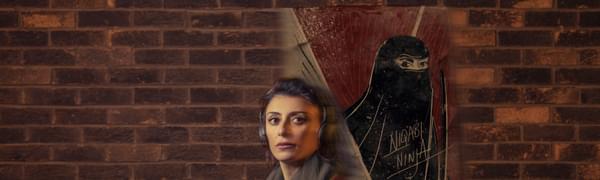 A woman wearing headphones stands facing the camera, in front of brick wall featuring a painting of a woman wearing a niqab.