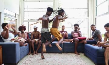A group of young people sit around on sofas as two young men dance in the middle