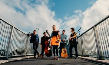 The Breabach members pose on a bridge with their instruments: fiddle, cello, bagpipes, guitar, and whistle