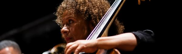 A close-up image of a member of the Chineke! Chamber Ensemble playing her cello, photographed from a low angle to show the position of her hand and bow.
