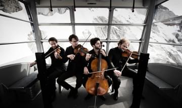 The Goldmund Quartet pictured playing their instruments seated close together in front of a window looking out on a snowy mountain scene.