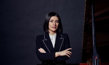 Mariam Batsashvilu pictured standing in front of a grand piano, looking into the camera with her arms folded.