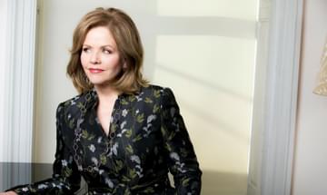 Renée Fleming, pictured leaning against a window and smiling as she gazes out, wearing a black silk shirt with a green and silver pattern.