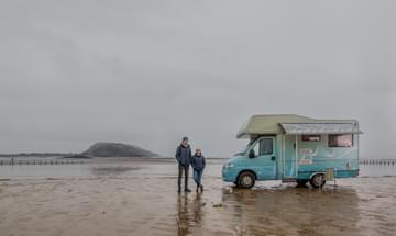 A man and a woman stand on a rainy beach next to a colourful caravan