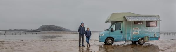 A man and a woman stand on a rainy beach next to a colourful caravan