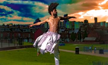 A man with dreadlocks wearing a skirt and trousers made of white fabric jumps in the air with his arms outstretched.
