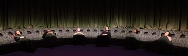 A group of people in a semicircle of reclined seating lie back with their eyes shut, with a large glowing light above them.