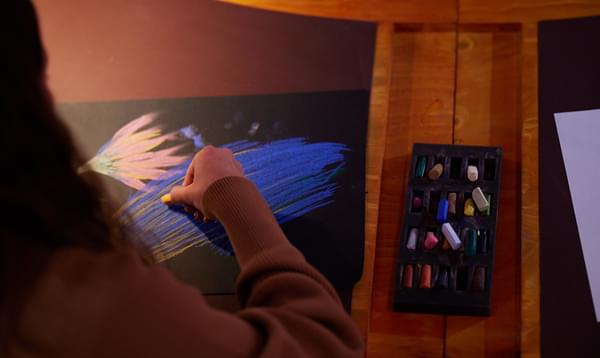 A close-up image of a person's hand holding a pastel crayon and sketching out blue shapes on a black piece of paper
