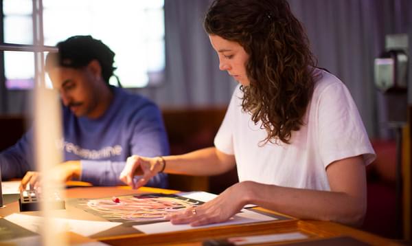 A woman in a white t-shirt and a man in a blue jumper sit at a desk drawing abstract shapes on black pieces of paper