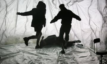 Three boys in silhouette against a translucent, crumpled background, with one of them lying on the floor and two of them standing over him.