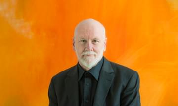 A bearded man in a black suit looks straight at the camera, sitting in front of a bright orange painting