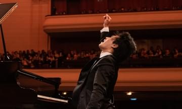 A young pianist sitting at the piano in the middle of a very passionate performance, with the right hand raised for extra drama. The artist is looking upwards and his face is sweaty and full of emotions.
