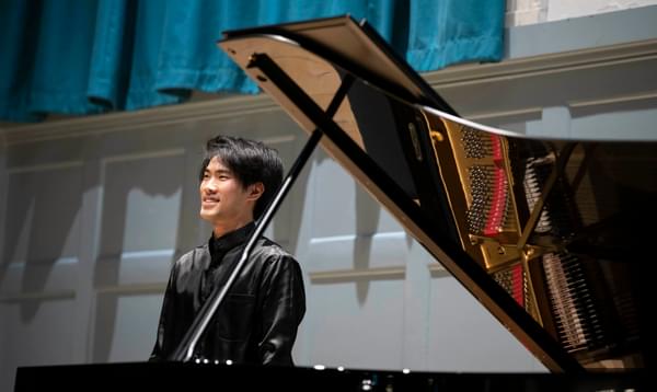 A man in a black shirt stands next to a grand piano smiling