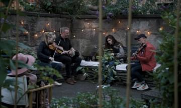 Four musicians sit in a leafy courtyard lit by fairylights playing their instruments.